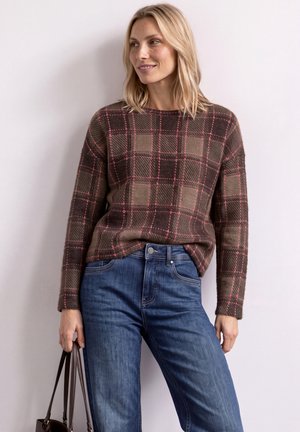 Lyshåret kvinde iført en brun ternet sweater med pink linjer og blå jeans, der holder en brun håndtaske og smiler let, foran en hvid væg.
