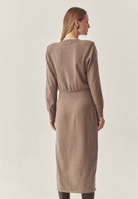 Adīta kleita mīkstā dūmainā brūnā krāsā, ar garām piedurknēm, piegulošu vidukli un midi garumu. Gluda faktūra ar aprocēm piedurknēs un apaļu kakla izgriezumu.