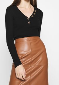 Haut noir côtelé à manches longues avec col en V et cinq boutons métalliques sur un côté, associé à une jupe crayon en cuir marron ajustée.