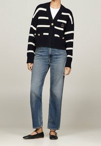 Cárdigan de rayas azul marino con rayas blancas, con escote en V, dos bolsillos delanteros y botones dorados; combinado con vaqueros azul claro y zapatos planos negros.