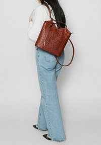 Borsa a secchiello in pelle marrone con perforazioni, tasca con zip, tracolla regolabile e superficie texture, indossata da una persona con jeans in denim chiaro.