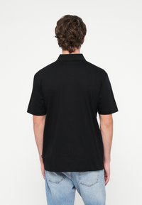 Melnas kokvilnas polo krekls ar apkakli, īsām piedurknēm un taisnu apakšu. Kreisajā pusē pie apakšas ir etiķete.