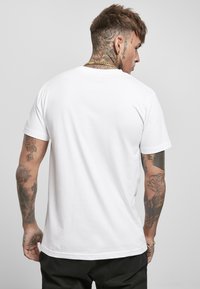 White cotton t-shirt with a crew neckline, short sleeves, and a plain design. Visible tattoos on arms, worn with black pants.