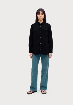 Femme debout portant une chemise noire boutonnée, un pantalon en velours côtelé bleu sarcelle et des tongs noires sur un fond clair uni.