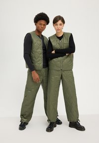 Gilets et pantalons en olive matelassés dans un matériau brillant, dotés d'une fermeture éclair et d'une finition texturée. Tops noirs à manches longues en dessous.