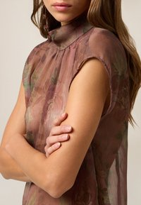 Blusa fluorescente in tessuto trasparente di color mauve con motivi floreali. Presenta un colletto alto e un design senza maniche, con una consistenza morbida e leggera.