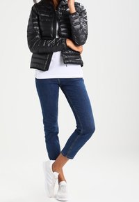 Sort dunjakke med lynlås foran og hætte, båret over en hvid skjorte. Blå jeans og hvide sneakers fuldender outfittet.