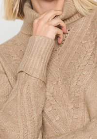 Beige pletený rolák se vzory z copánků a žebrovanými manžetami, s texturovanou tkaninou a volným střihem.