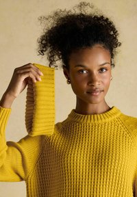 Frau mit lockigem Haar, die einen strukturierten gelben Pullover trägt, hält ein passendes gelbes Strickhaarband vor einem schlichten beigen Hintergrund.