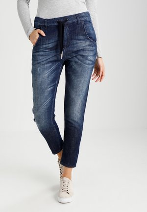 Femme portant un pantalon de jogging en denim bleu foncé avec taille à cordon et des baskets blanches, main dans la poche, debout devant un fond uni.