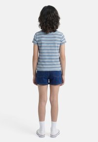 Παιδί με σγουρά μαλλιά που φοράει μπλε ριγέ μπλούζα, τζιν σορτς, λευκές κάλτσες και λευκά αθλητικά παπούτσια, στέκεται με την πλάτη γυρισμένη σε λευκό φόντο.