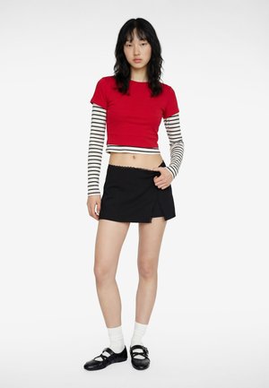 Jeune femme debout portant un top court rouge sur un haut à manches longues rayé noir et blanc, une mini-jupe noire, des chaussettes blanches et des chaussures noires.