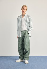 Stribet lyseblå og hvid skjorte med knapper over en hvid tanktop, parret med løse grønne cargo-bukser og hvide sneakers.