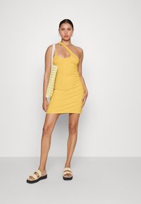 Vestido mini amarelo, sem mangas, com decote assimétrico e design justo. Combinado com sandálias de creme com babados e uma bolsa de ombro de palha.