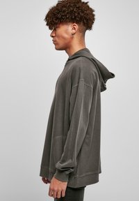 Stort grå hettegenser med snøring i hetten, ribbede mansjetter og hem, samt mykt, strukturert stoff vist på en modell i profil.