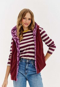 Młoda kobieta z falującymi blond włosami, ubrana w bordo w paski koszulę z długim rękawem, bordo kamizelkę w kwiaty i niebieskie dżinsy, lekko się uśmiecha.