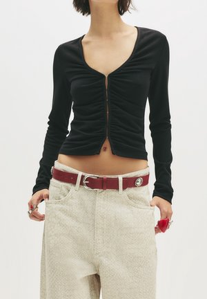 Femme portant un crop top noir froncé à manches longues, un pantalon taille haute léger texturé, une ceinture rouge et des bagues aux deux mains.