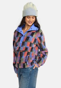 Farverig fleece-pullover med patchwork-design i rød, grøn, blå og sort. Har en blå krave, lomme og ribbet hue.