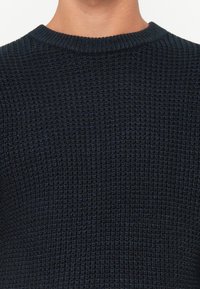 Primo piano di una persona che indossa un maglione a maglia strutturata di un blu scuro con girocollo a coste.