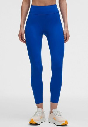 Leggings i ensfarvet klar blå med høj talje, båret med hvide og orange atletik sko på en stående person.