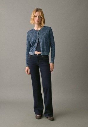 Giovane donna che indossa jeans blu scuri, un top corto scuro e un cardigan blu con bottoni, appoggiata su uno sfondo grigio uniforme.