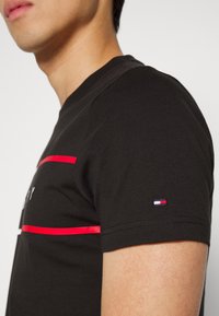 Camiseta de algodón negra con mangas cortas, que presenta rayas horizontales rojas y un pequeño parche de logo tricolor en la manga.
