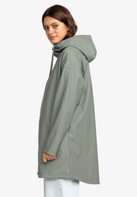Mujer vestida con un chubasquero verde salvia con capucha, cremallera frontal y logo en la manga, combinado con jeans azules claros, mirando de lado.