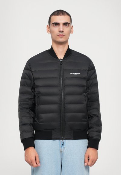 Black padded bomber jacket with a zip front, ribbed cuffs, and waist. Features a small logo on the left chest, smooth texture.