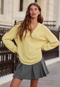 Dzeltens V-veida kakla džemperis ar garām piedurknēm, kombinēts ar pelēku kroku svārku ar pundurkoka rakstu. Mīksta tekstūra, brīvs piegriezums.