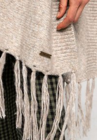 Poncho de punto beige claro con flecos largos y una pequeña etiqueta metálica. Al fondo se puede ver una tela cuadros en verde.