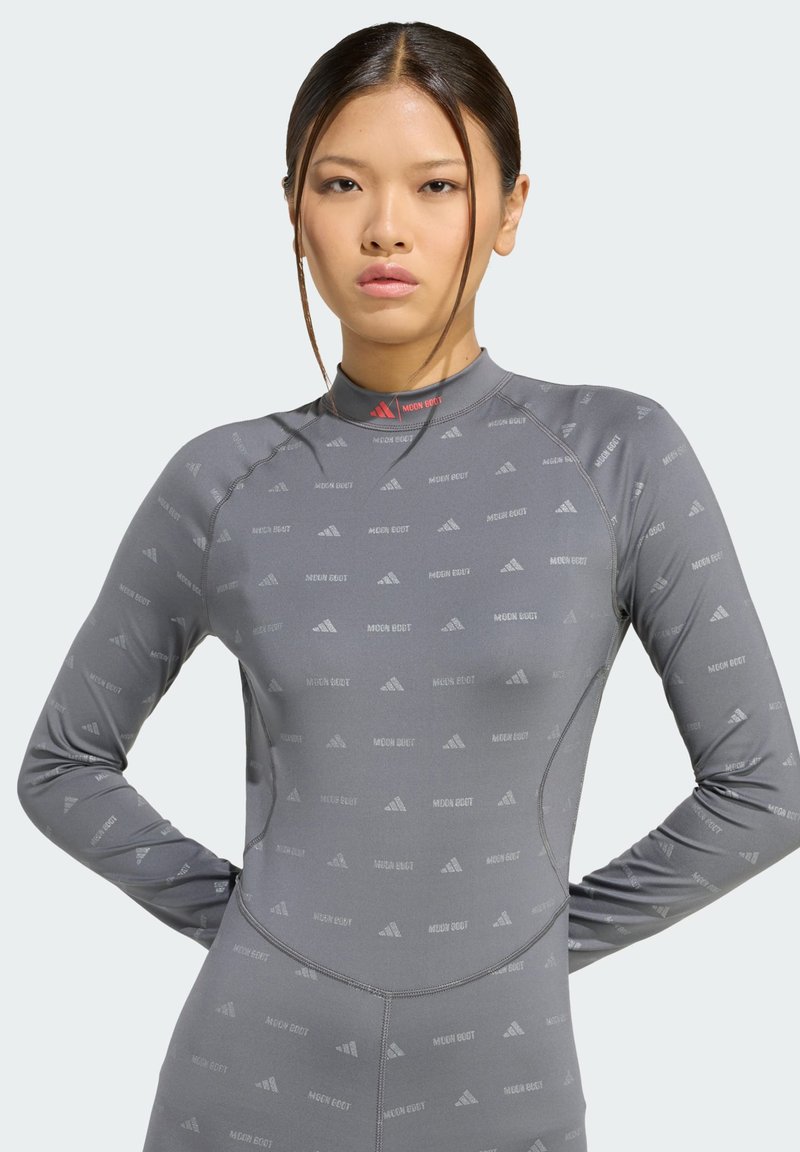 Jonge vrouw met steil haar, gekleed in een aansluitend grijs langemouwen shirt met herhaalde "MOON BOOT" en logo patronen, staande met handen op de heupen.