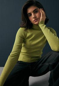 Rippstrick-Turtleneck-Pullover mit langen Ärmeln in leuchtendem Grün, kombiniert mit dunklen Jeans. Der Stoff hat eine strukturierte Optik und eine figurbetonte Silhouette.