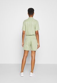 Personne debout, de dos, portant une chemise à manches courtes vert clair et un short assorti, avec des baskets blanches, sur fond uni.