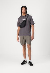 Gray t-shirt with logo, paired with tan checked shorts. Black waist bag worn crossbody, black sneakers, and white socks. Minimalist design.