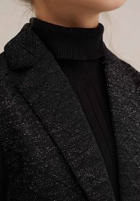 Svart texturerad kavaj med en subtil lyster, lager på lager över en svart ribbad polotröja. Fokusera på tygdetaljerna och formen på kragen.