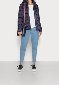 Giacca puffer blu navy con motivo a righe orizzontali, t-shirt bianca, jeans azzurri chiari e sneakers blu navy. Borsa a tracolla con una tracolla a righe.