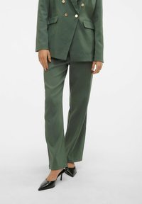 Femme portant un costume vert foncé sur mesure avec des boutons dorés et des talons aiguilles noirs pointus, debout devant un fond blanc uni.