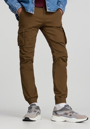 Uomo che indossa pantaloni cargo marroni, calzini bianchi, sneaker grigie e beige, con le mani nelle tasche e una giacca di jeans.