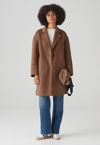 Brown wool coat with a notched collar, single button closure, and wide sleeves, paired with light blue jeans and white sneakers.