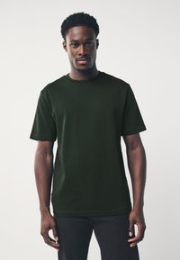 Camiseta de algodón verde oscuro con cuello redondo, mangas cortas y un corte holgado. Textura suave y detalles de costura mínimos.