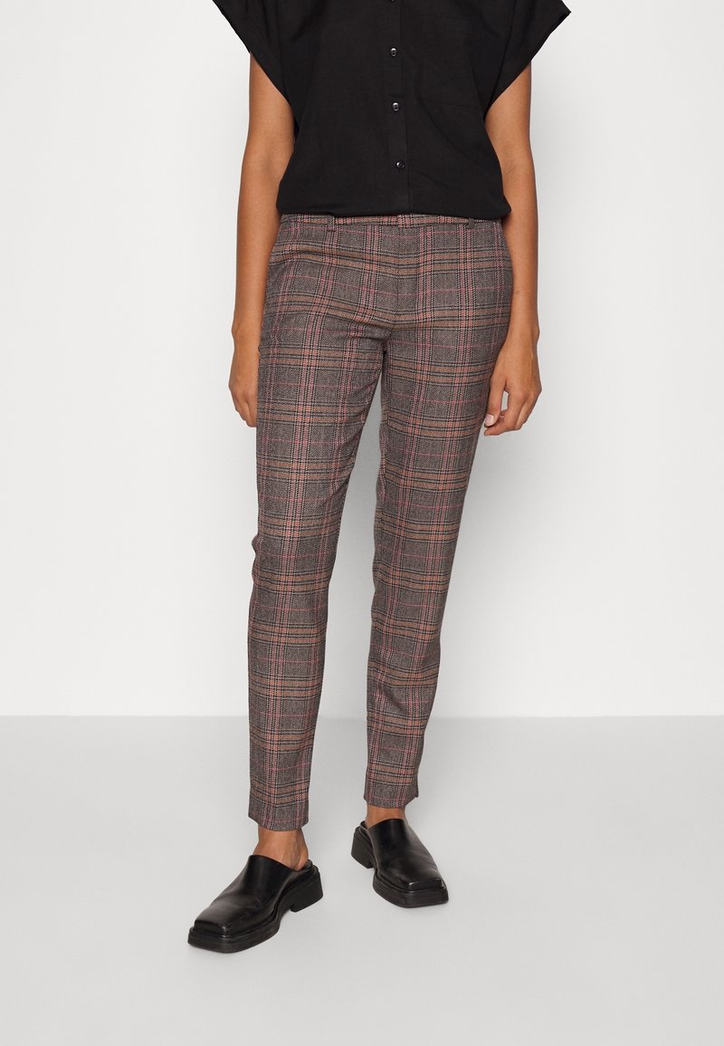 Personne portant un pantalon slim à carreaux gris avec des lignes rouges, associé à une chemise noire boutonnée et des chaussures noires slip-on, debout sur un sol gris.