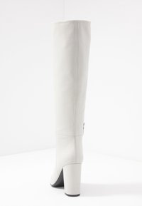 Bota de cuero gris hasta la rodilla con una textura suave, tacón cuadrado y suela negra. Presenta una costura en la parte trasera y un detalle lateral.