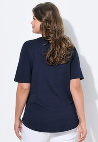 Nainen, jolla on pitkät laineikkaat ruskeat hiukset, pukeutuneena tummansiniseen lyhythihaiseen puseroon ja valkoisiin housuihin, seisoo sivukamarissa yksinkertaista valkoista taustaa vasten.