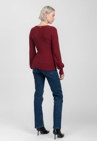 Bordeauxgekleurde gebreide trui met lange pofmouwen en een ronde halslijn, gecombineerd met blauwe jeans en zwarte hoge hakken.