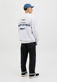 Sudadera gris con la impresión "Atletismo Funcional", jeans de mezclilla oscura con dobladillos enrollados y zapatillas deportivas negras con suelas blancas, complementadas con una gorra azul.