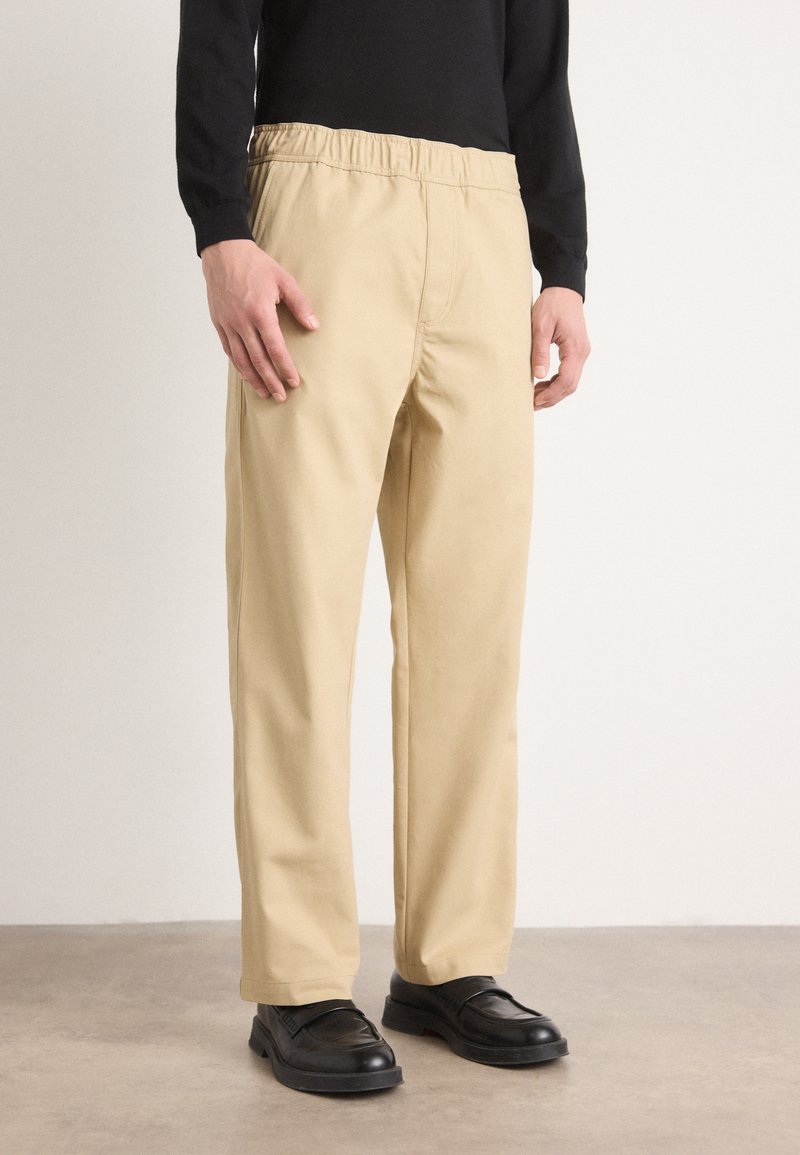 Pantalons beiges avec une taille élastique, design à jambe droite et texture lisse. Portés avec des chaussures noires sur un fond neutre.