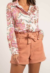 Blouse à manches longues avec un fond blanc et un motif floral paisley coloré. Accompagnée de shorts ceinturés marron clair avec des ourlets roulés.