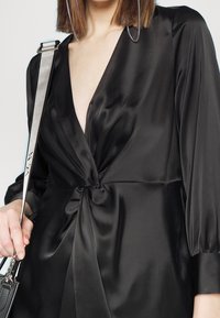 Femme portant une robe en satin noire avec un décolleté en V profond et un détail de nœud torsadé, tenant un sac avec une sangle grise sur l'épaule.