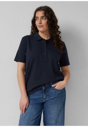 Frau mit lockigem Haar, die ein marineblaues Poloshirt mit kurzen Ärmeln und blaue Jeans trägt, steht mit einer Hand in der Tasche und schaut zur Seite.