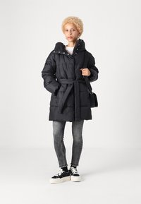 Giacca a vento nera imbottita con vita regolabile da una cintura, cappuccio e chiusura a bottoni a pressione. Abbinata a jeans skinny grigi e sneakers nere. Texture liscia e isolante.